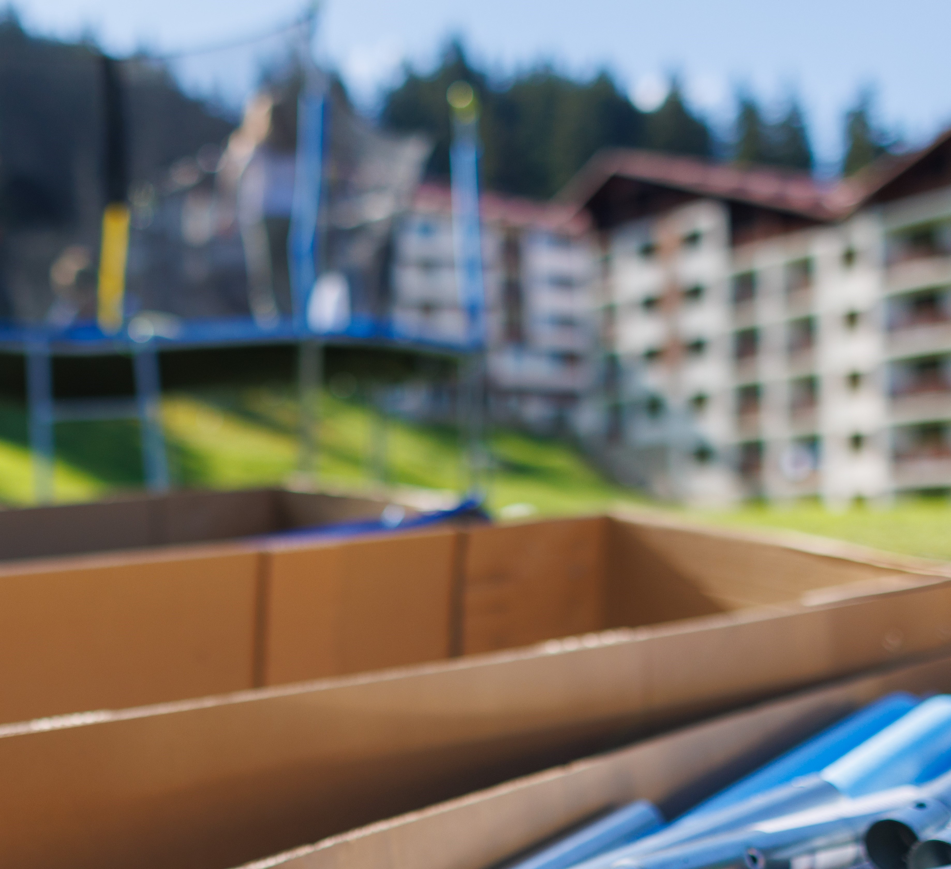 Large cardboard boxes with iron beams and other kit to assemble a netted trampoline as in the background next to people assembling a trampoline on a backyard lawn